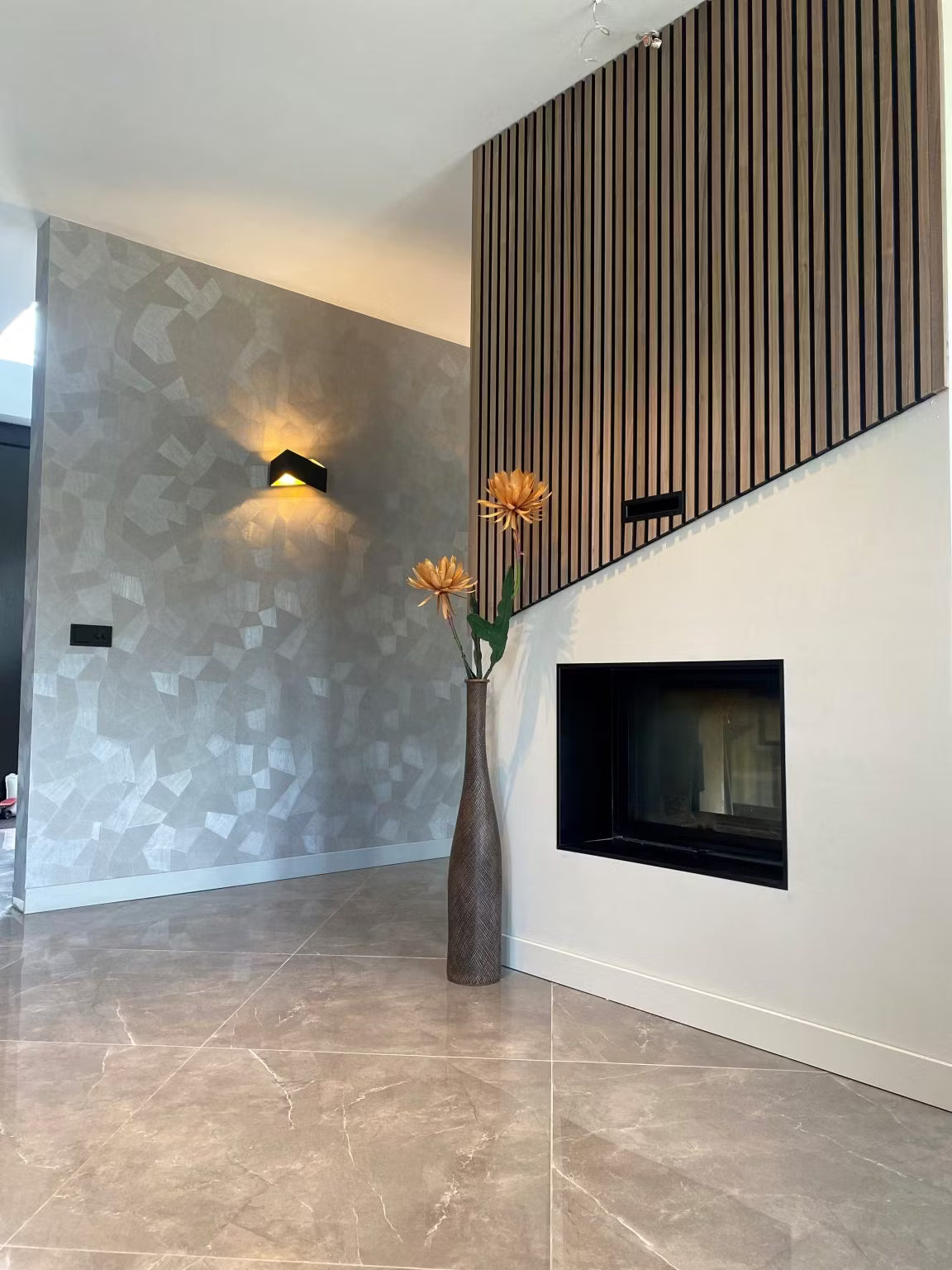 Modern living room with geometric-patterned wallpaper, Akupanel wood acoustic paneling, wall-mounted fireplace, and a tall vase of orange flowers.